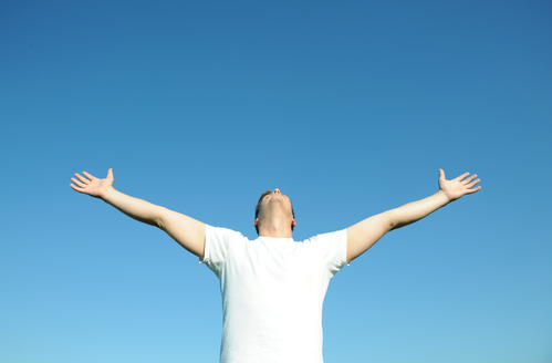 Young man in white t-shirt arms open, blue sky. Above space for text or graphics.